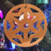 A handmade wooden Christmas tree ornament, circular in shape, crafted from light brown wood with a natural grain pattern. It features an intricate, symmetrical cutout design resembling a star or snowflake, with multiple pointed shapes radiating from a central hole. The ornament is attached to a small metal hook for hanging, showcasing its rustic and festive craftsmanship.