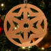 A handmade wooden Christmas tree ornament, circular in shape, crafted from light brown wood with a natural grain pattern. It features an intricate, symmetrical cutout design resembling a star or snowflake, with multiple pointed shapes radiating from a central hole. The ornament is attached to a small metal hook for hanging, showcasing its rustic and festive craftsmanship.