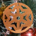 A handmade wooden Christmas tree ornament, circular in shape, crafted from light brown wood with a natural grain pattern. It features an intricate, symmetrical cutout design resembling a star or snowflake, with multiple pointed shapes radiating from a central hole. The ornament is attached to a small metal hook for hanging, showcasing its rustic and festive craftsmanship.